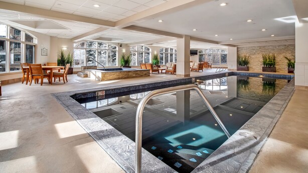 A spacious indoor pool area with a hot tub, seating, and large windows offering views of the outdoors.