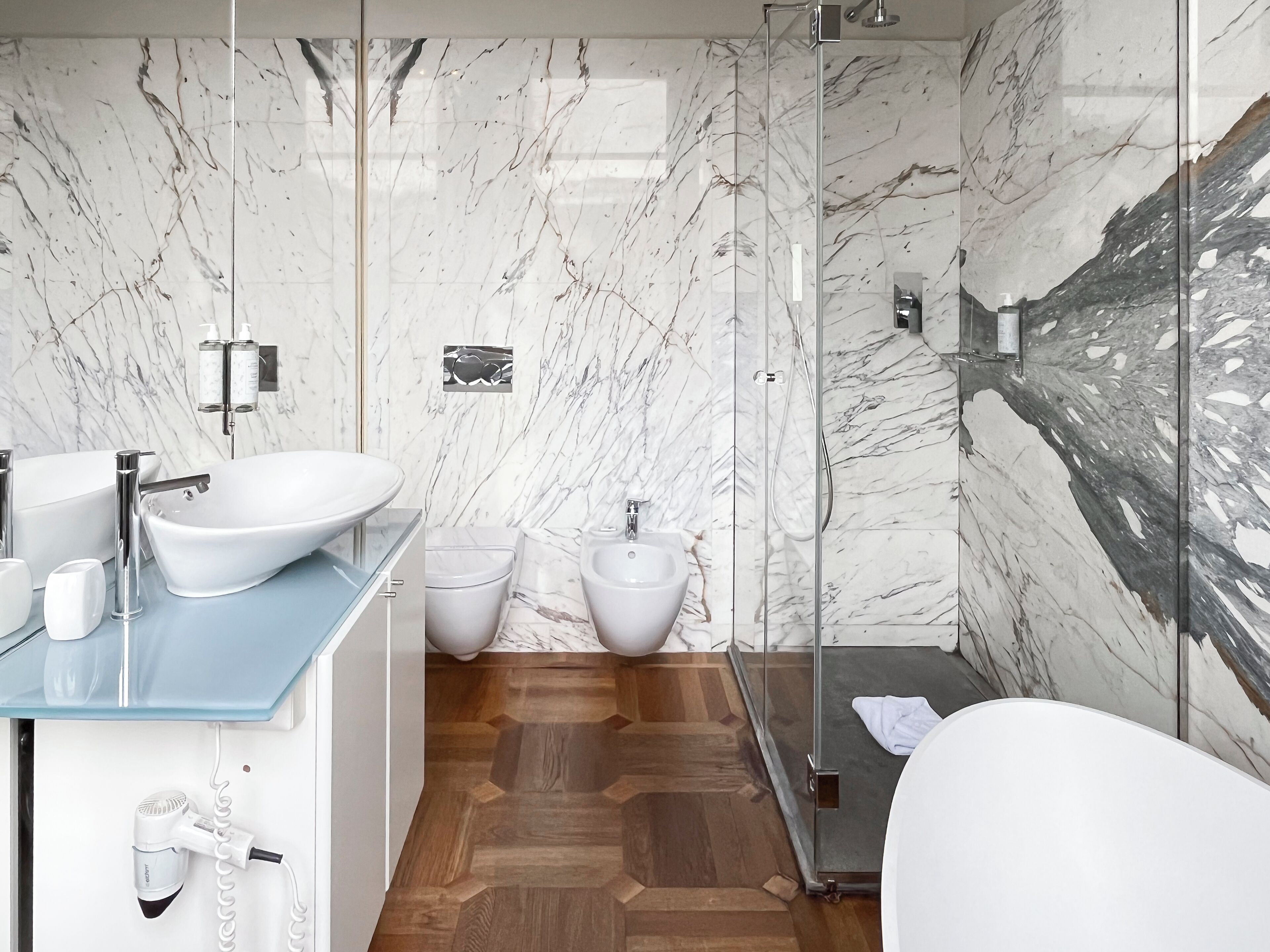 A modern bathroom with a marble backsplash, a freestanding bathtub, a glass shower enclosure, and a wooden floor.