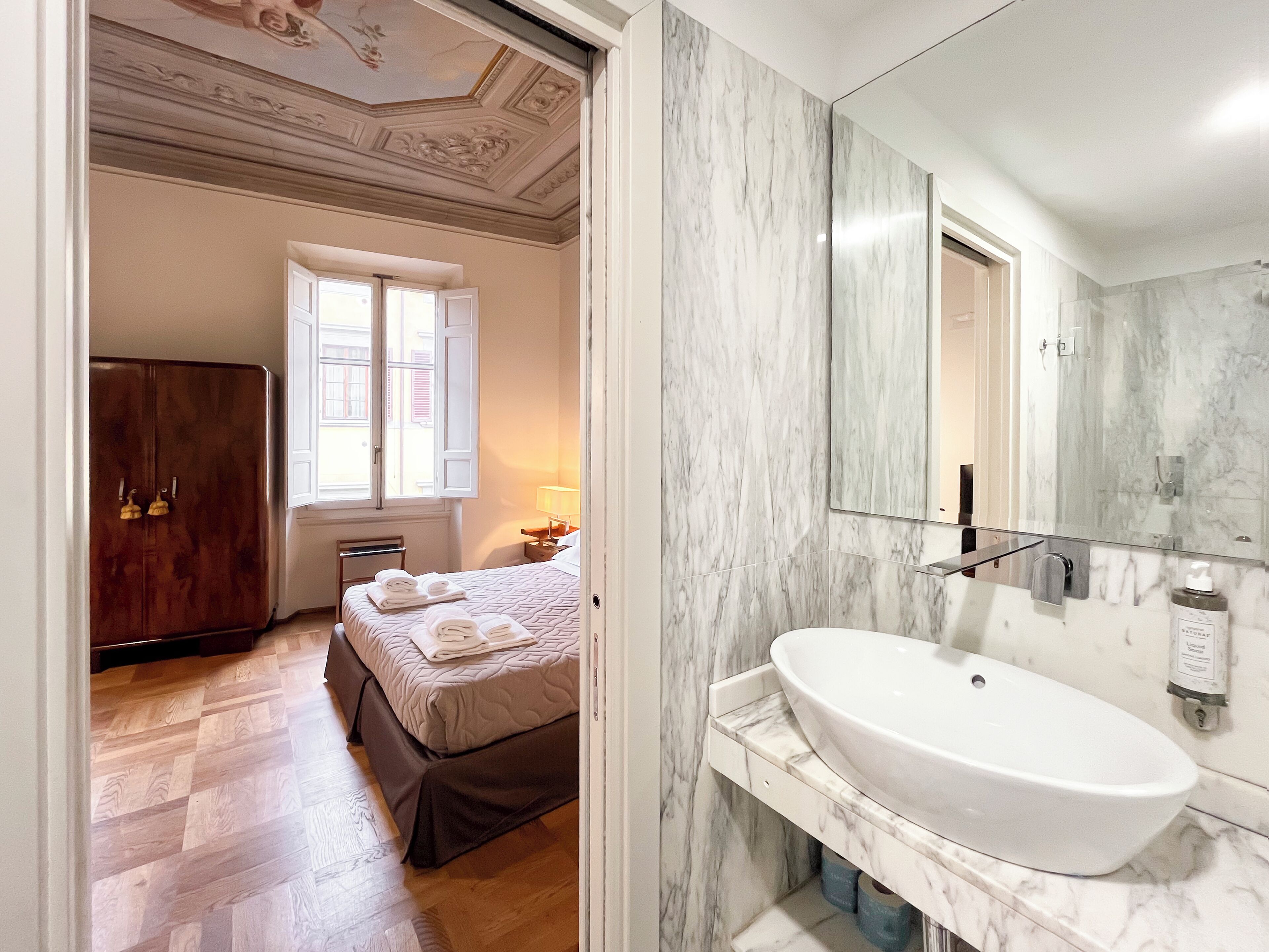 Une chambre d’hôtel comprenant un lit, une armoire, une salle de bain avec un grand lavabo et un miroir, ainsi qu’une fenêtre avec des volets.