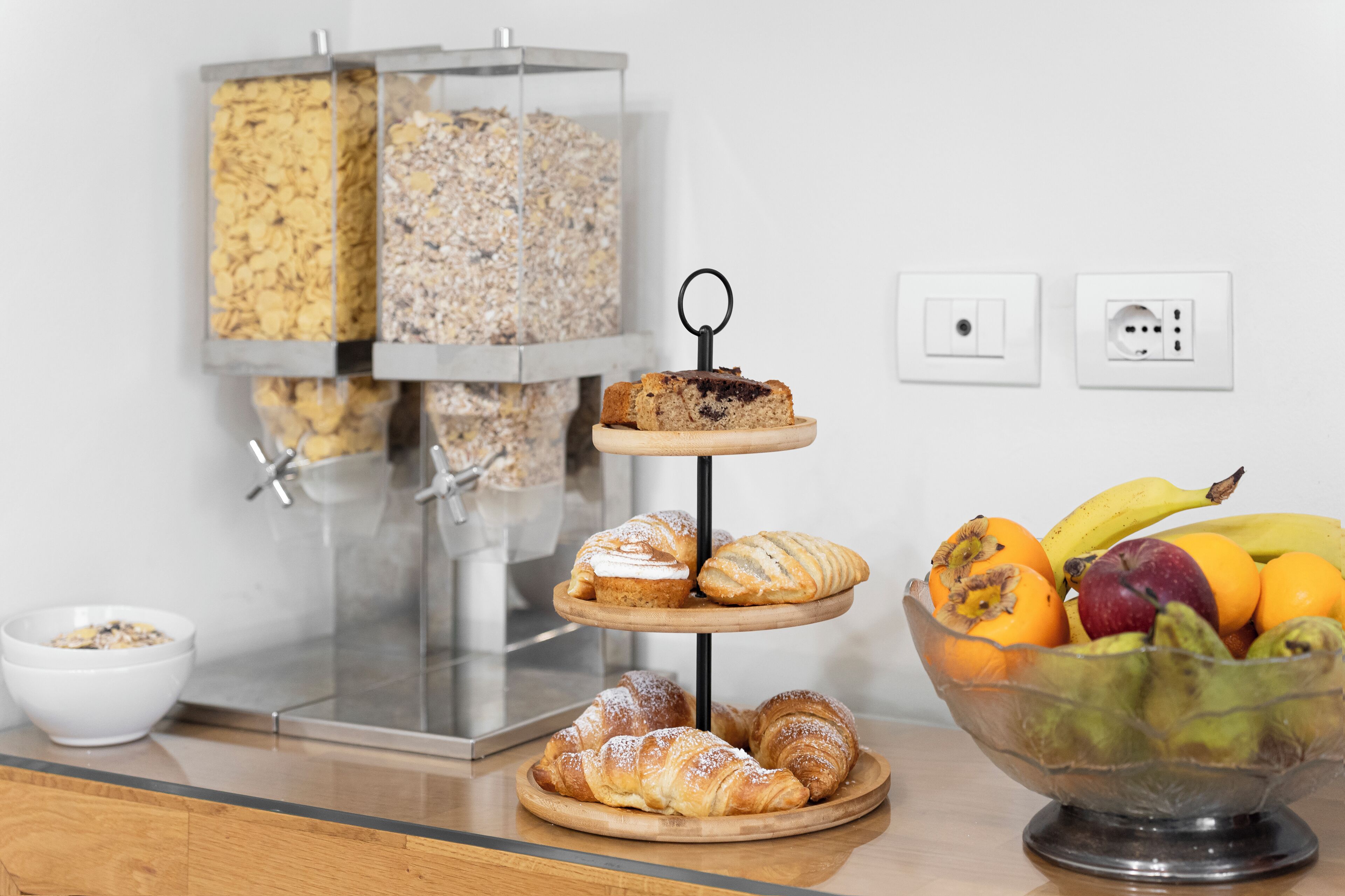 A breakfast setup with a three-tiered stand of pastries, a bowl of assorted fruits, and a container of cereal flakes.