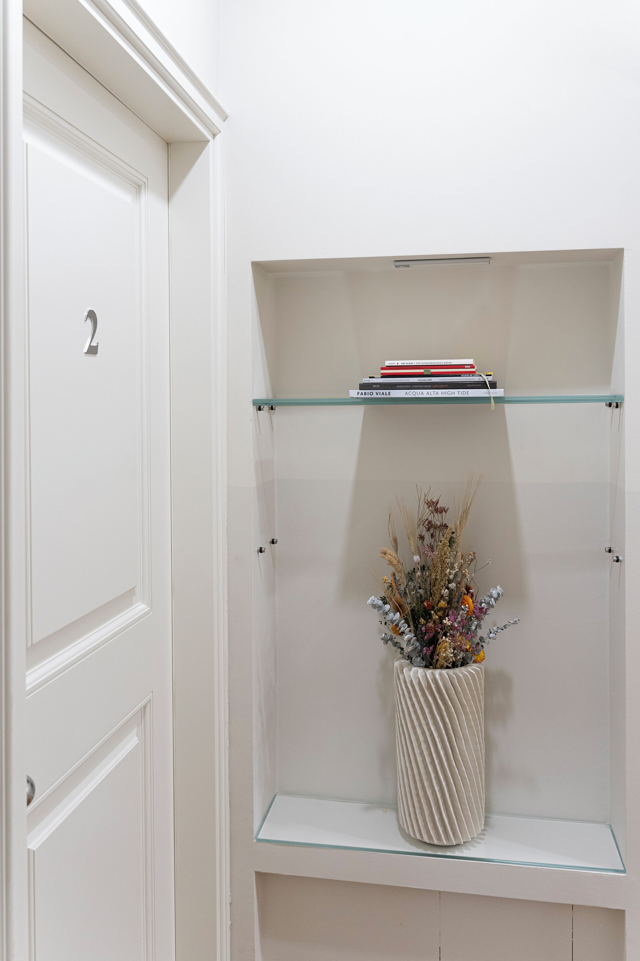 A white room with a glass shelf holding books and a vase with dried flowers.