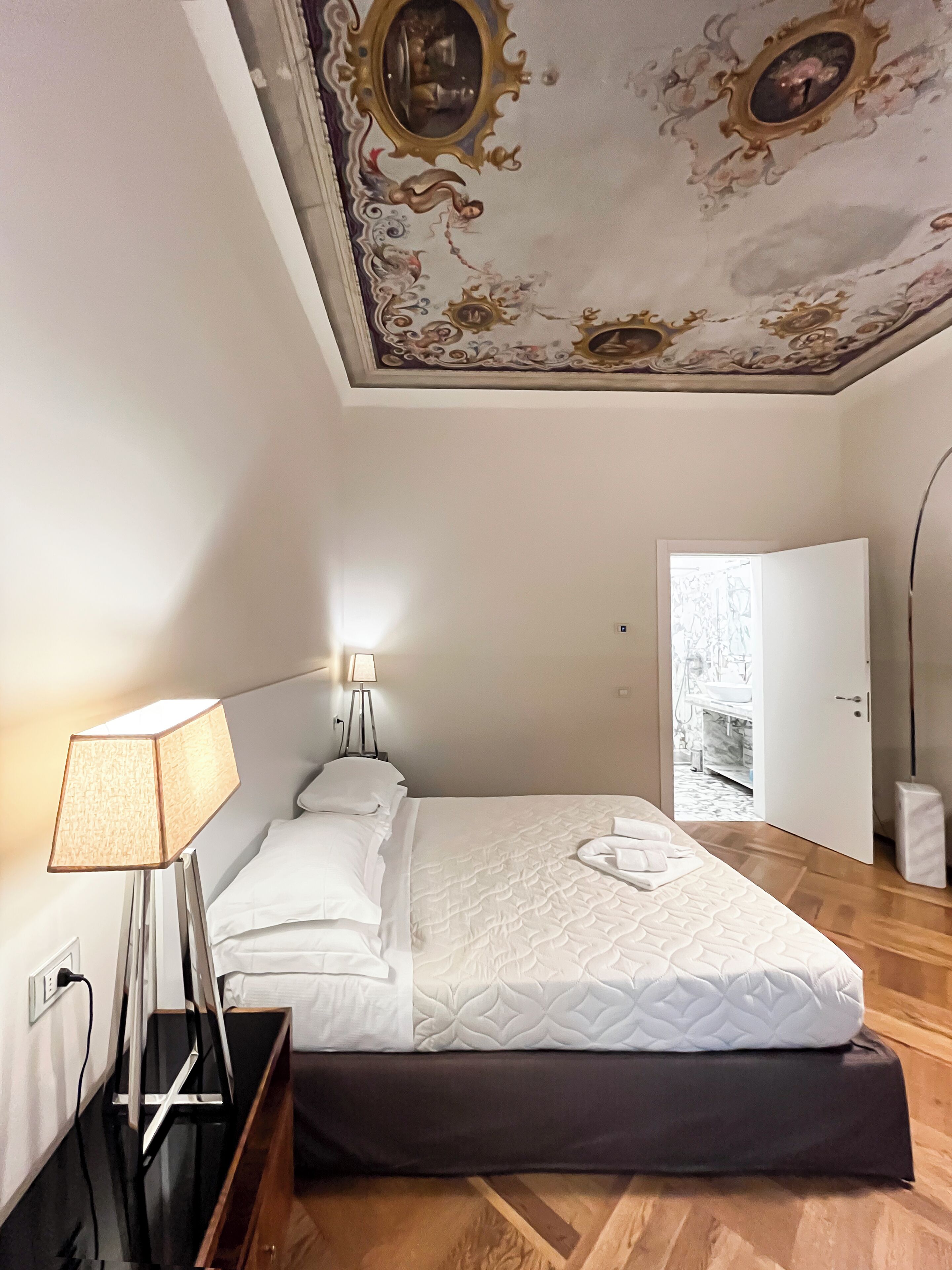 A bedroom with a bed, bedside lamps, a wooden floor, and a ceiling with ornate designs.