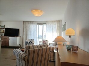 Un salon équipé d’un téléviseur à écran plat, d’un canapé, d’une table à manger avec une lampe et d’une fenêtre avec des rideaux.