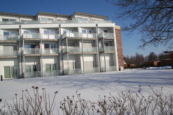 A multi-story residential building with balconies, a brick section, and a snow-covered ground.