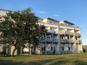 A multi-story residential building with balconies, a tree in the foreground, and a clear sky.