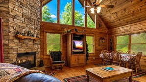L’intérieur d’une cabane en rondins, avec une cheminée, un téléviseur, un coin repas et une grande fenêtre donnant sur la forêt.