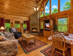 A cozy cabin interior with a fireplace, wooden furniture, and large windows offering a view of the outdoors. - Cub Crossing, Free Attraction Tickets, Hot Tub, Pool Table, WIFI!!! (Sevierville)