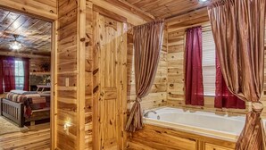 A log cabin interior with a wooden floor, a large bathtub, and a bed with a red blanket.