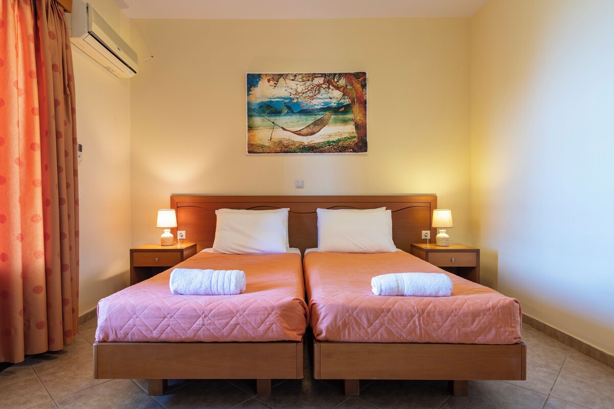 A hotel room with two beds, each with a pillow and a towel, a painting of a beach scene on the wall, and an air conditioning unit above the beds.