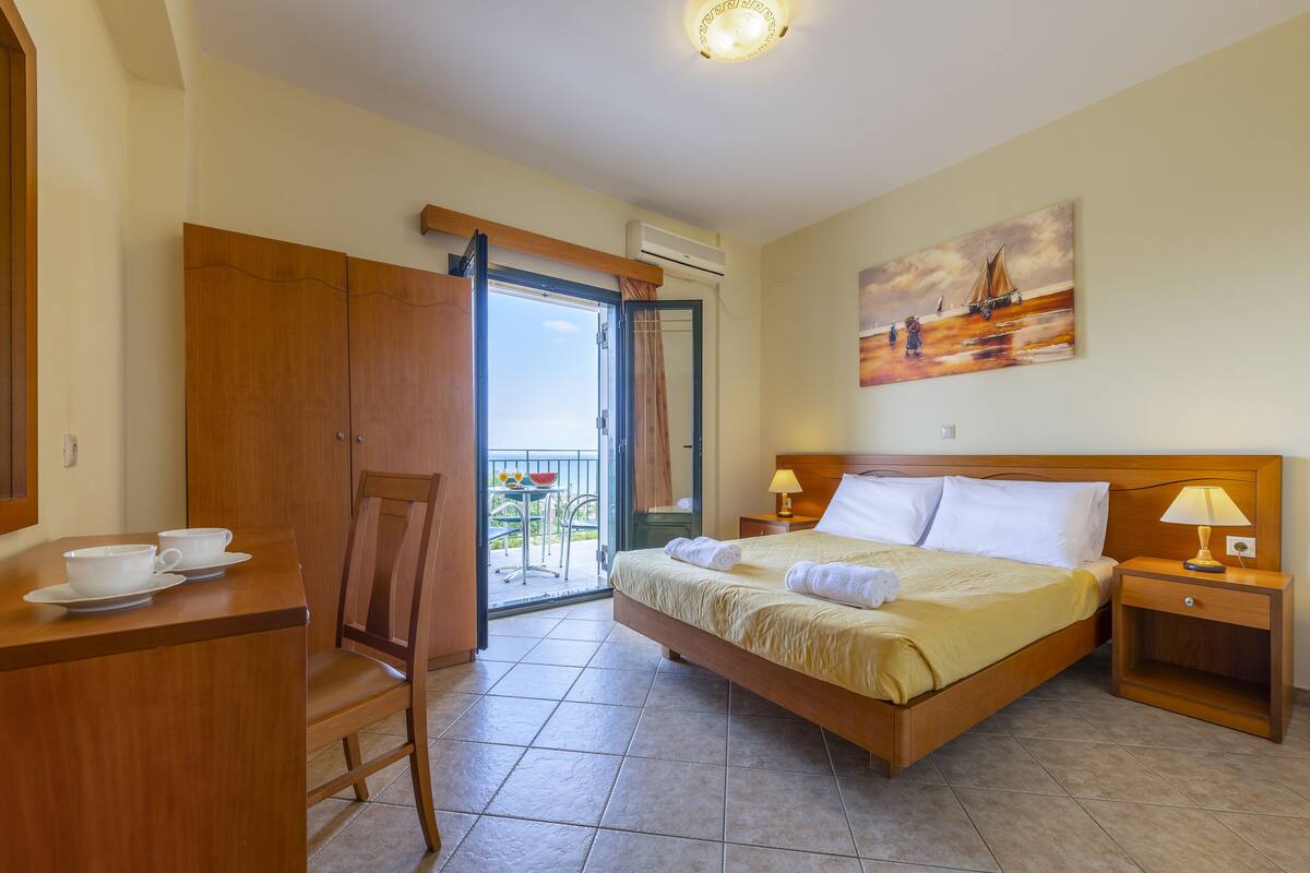Ein Hotelzimmer mit einem Bett, einem Schreibtisch mit Tassen, einem Stuhl, einem Balkon mit Tisch und Stühlen sowie einem Bild an der Wand.