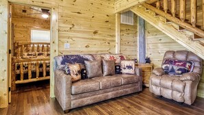 A log cabin interior with a wooden staircase, a leather sofa with animal-themed cushions, and a plush armchair. - Forever Yours, 2 Hot tubs, FREE Attraction tickets, WIFI, Near The Parkway!!! (Pigeon Forge)