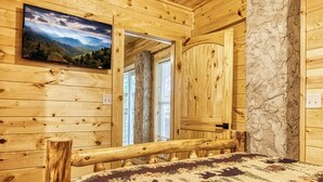 A wooden cabin interior with a bed, a mounted TV, and a door leading to a balcony. - Forever Yours, 2 Hot tubs, FREE Attraction tickets, WIFI, Near The Parkway!!! (Pigeon Forge)