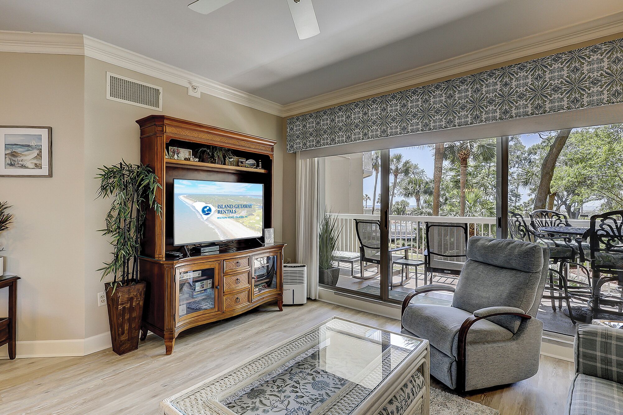 A living room with a television displaying a rental advertisement, a glass coffee table, a grey armchair, and a view of the outdoors through large windows.