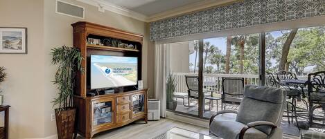 A living room with a television displaying a rental advertisement, a glass coffee table, a grey armchair, and a view of the outdoors through large windows.