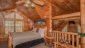 A log cabin bedroom with a bed, a wooden bench with teddy bears, a ceiling fan, and a window with a view of trees. - Bearskin Romance- Free Attraction Tickets ,WIFI, Pool Table, Air Hockey, Hot Tub (Sevierville)