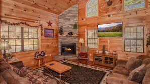 A log cabin interior with a fireplace, leather sofas, a wooden coffee table, and large windows. - Bearskin Romance- Free Attraction Tickets ,WIFI, Pool Table, Air Hockey, Hot Tub (Sevierville)