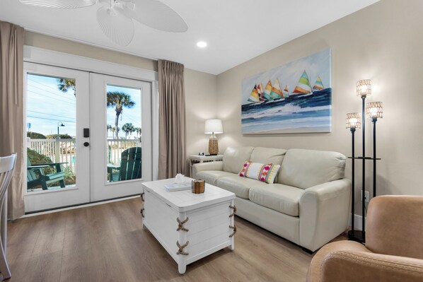 A living room with a white sofa, a white coffee table, a painting of sailboats, a floor lamp, and a view through a sliding door to the outside. - Emerald Gulf, Pool & Hot tub at your patio,-FREE BEACH ITEMS - pocket doors (Miramar Beach)