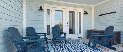 A covered porch with three Adirondack chairs, a small table, and a patterned rug.