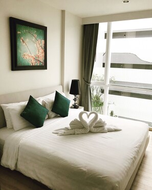 A hotel room with a neatly made bed, a framed picture on the wall, a lamp, and a view of the outside through the window. - The Base Downtown Condo by Joy (Wichit)