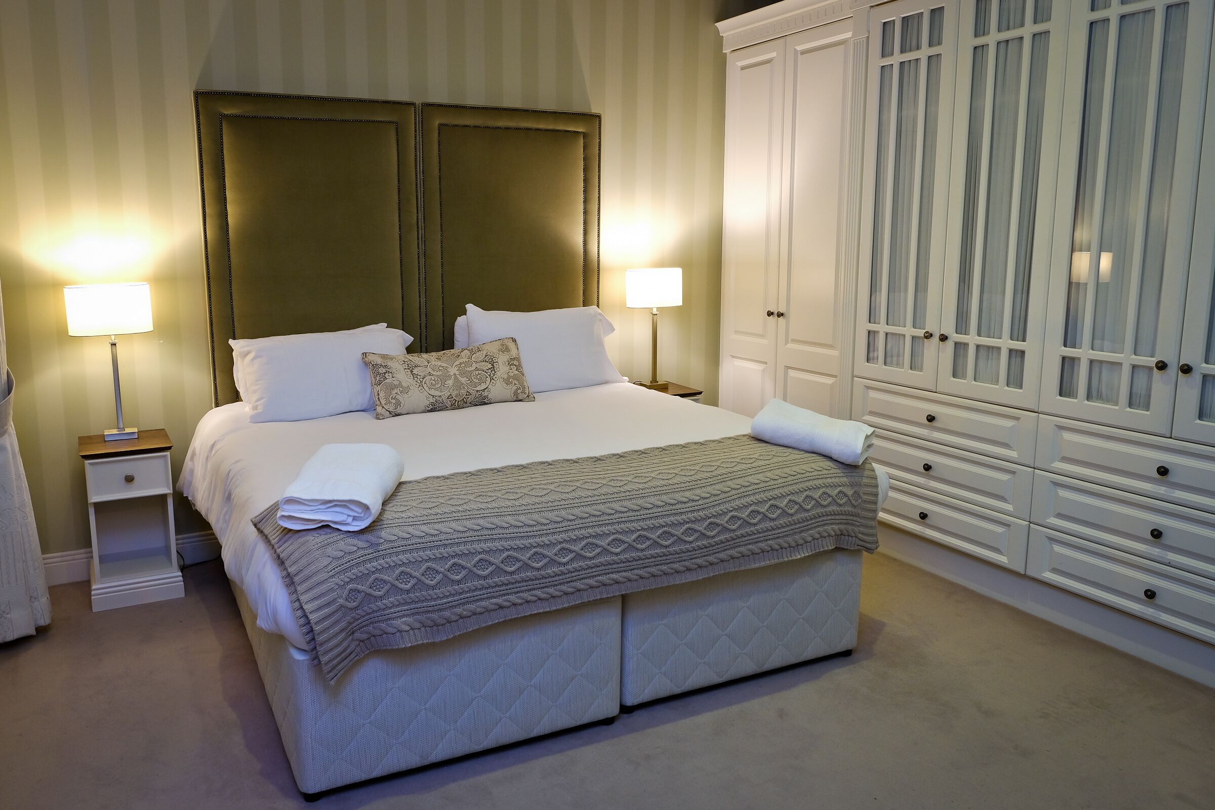 A neatly made bed with a quilted cover, two bedside lamps, a nightstand, and a large white wardrobe with multiple drawers.