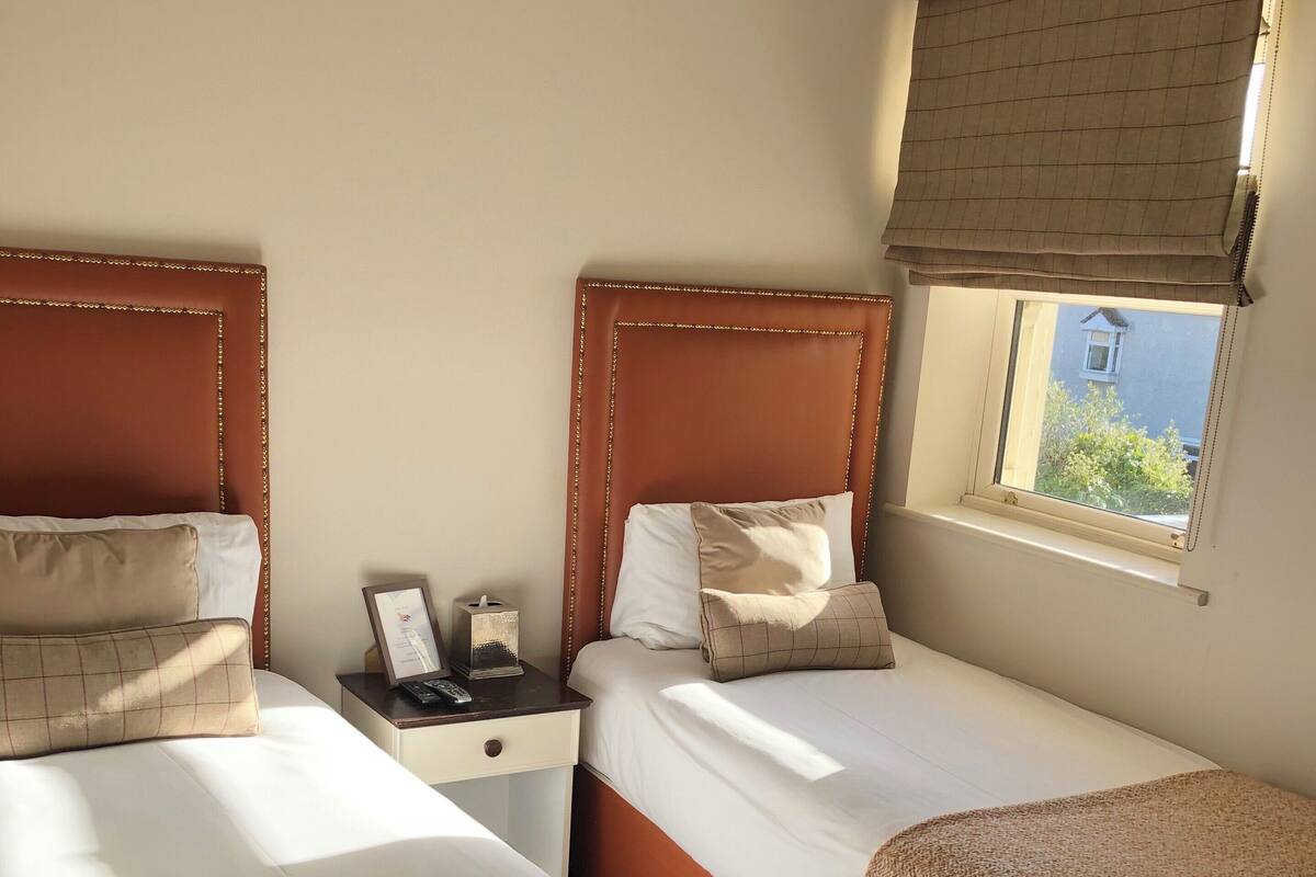 Quarto de hotel com duas camas de solteiro, uma mesinha de cabeceira com telefone e abajur, uma janela com vista para áreas verdes e cabeceira marrom.