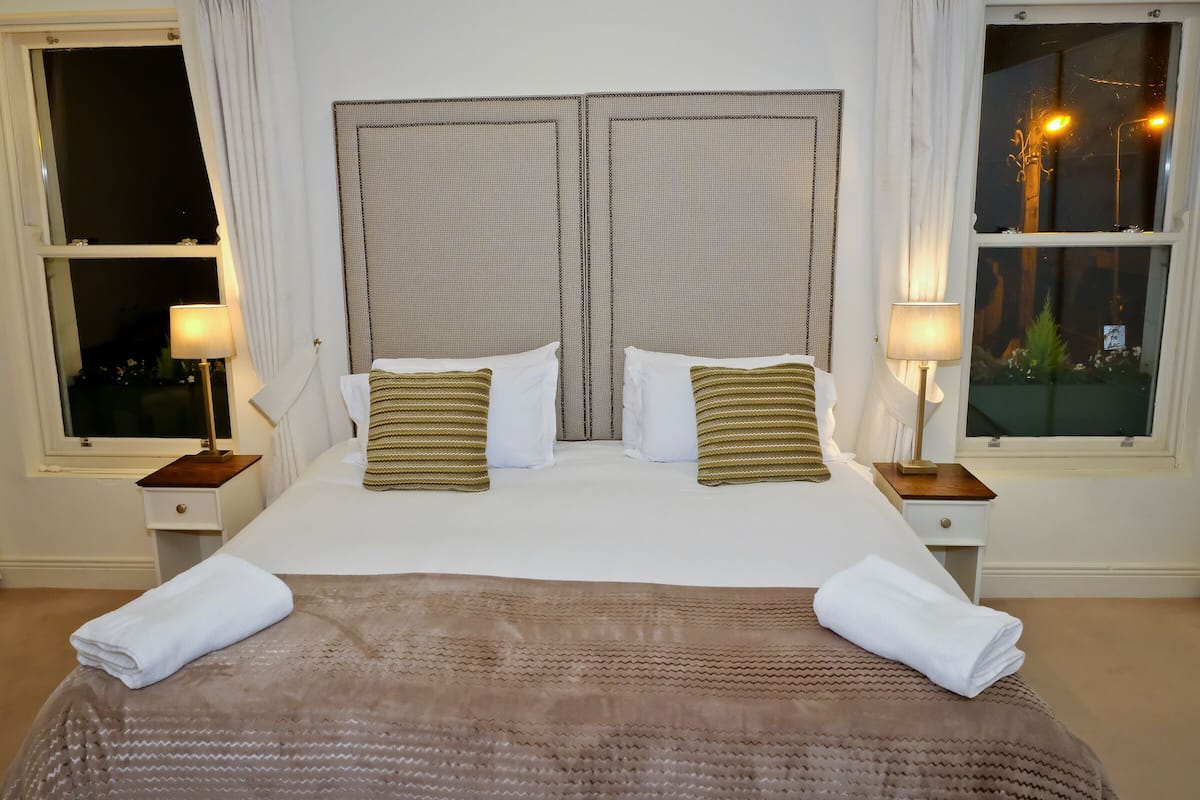 Uma cama de casal com um cobertor marrom, duas almofadas brancas e duas almofadas listradas. Há duas mesas de cabeceira com luminárias, uma janela com cortinas e vista para árvores do lado de fora.
