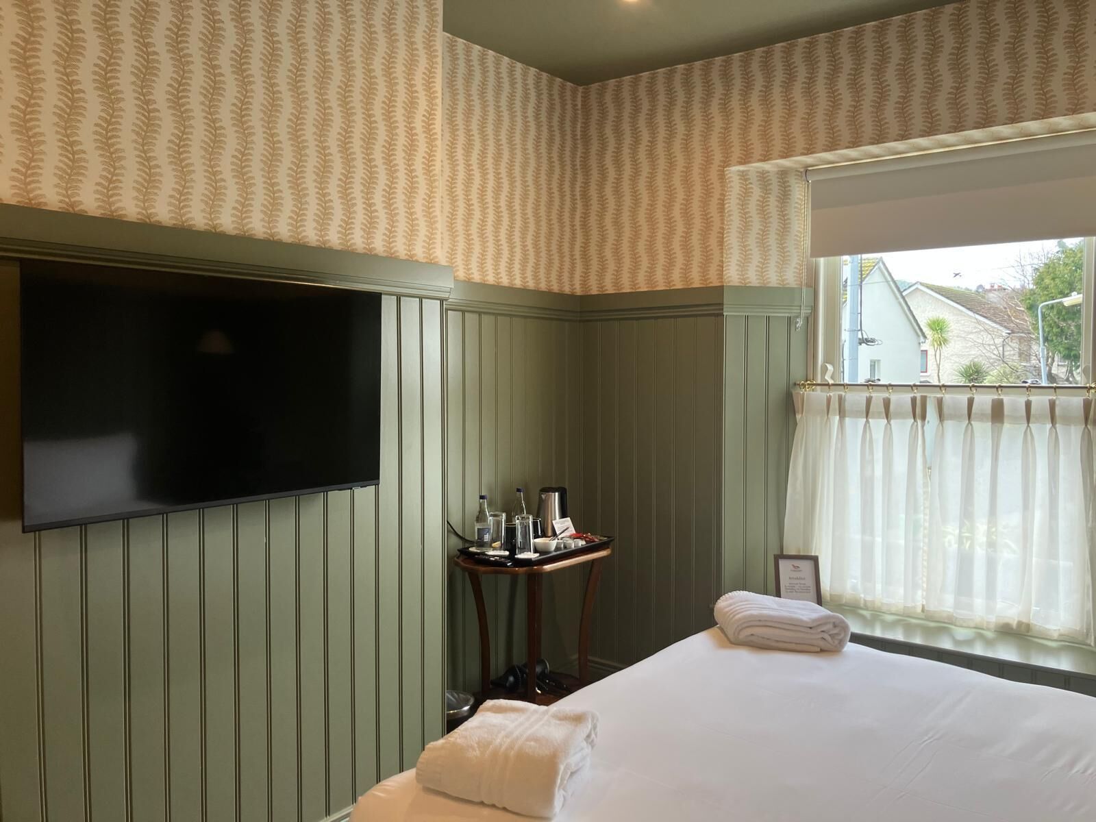 A hotel room with a bed, a TV mounted on the wall, a small table with toiletries, and a window with curtains.