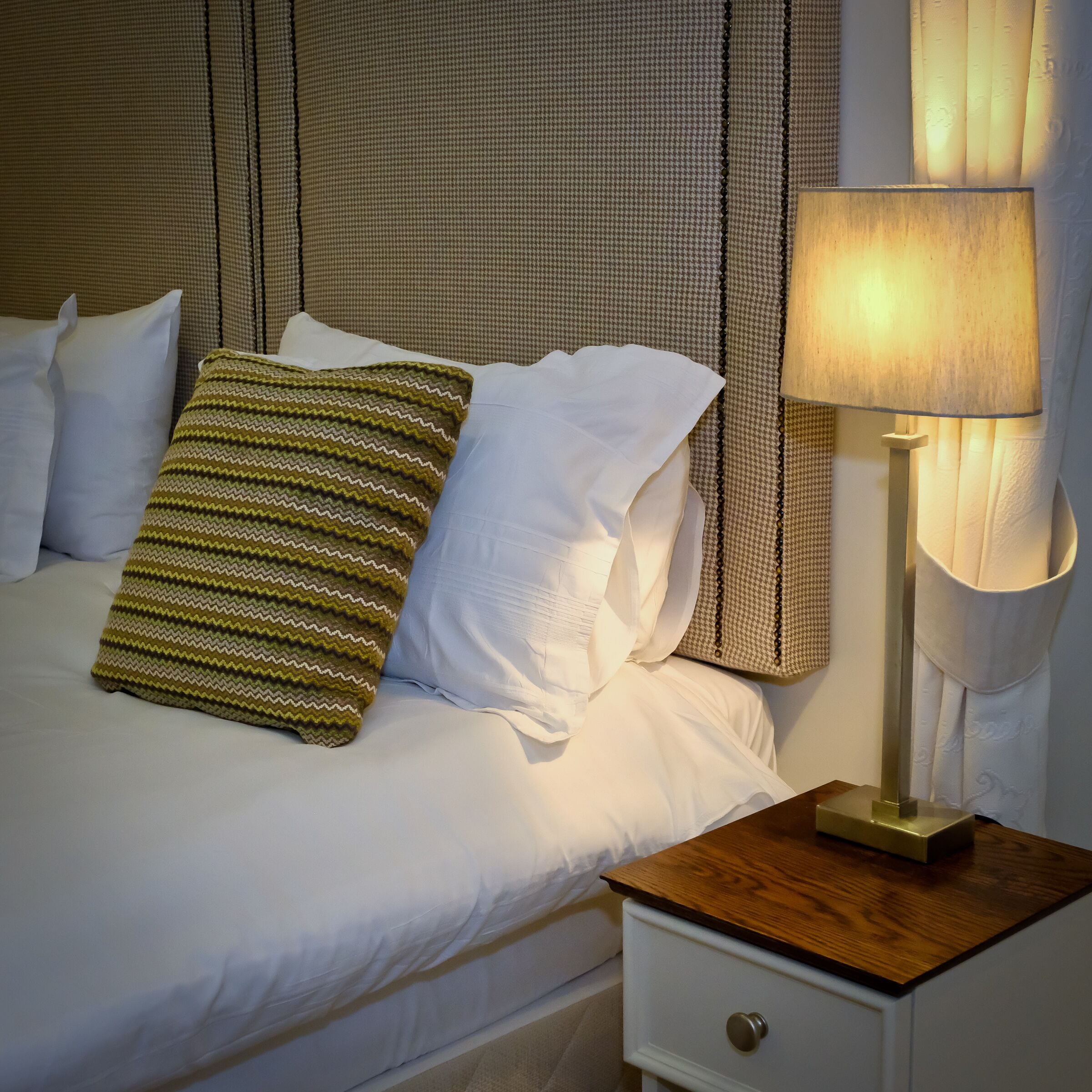 A neatly made bed with white linens and a green striped pillow, a bedside table with a lamp, and a houndstooth-patterned curtain.