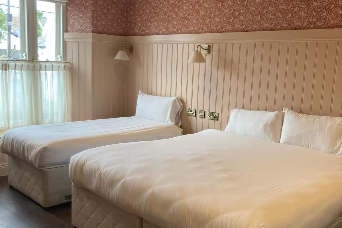 Quarto de hotel com duas camas, uma janela com cortinas, parede com linhas verticais e uma luminária no teto.