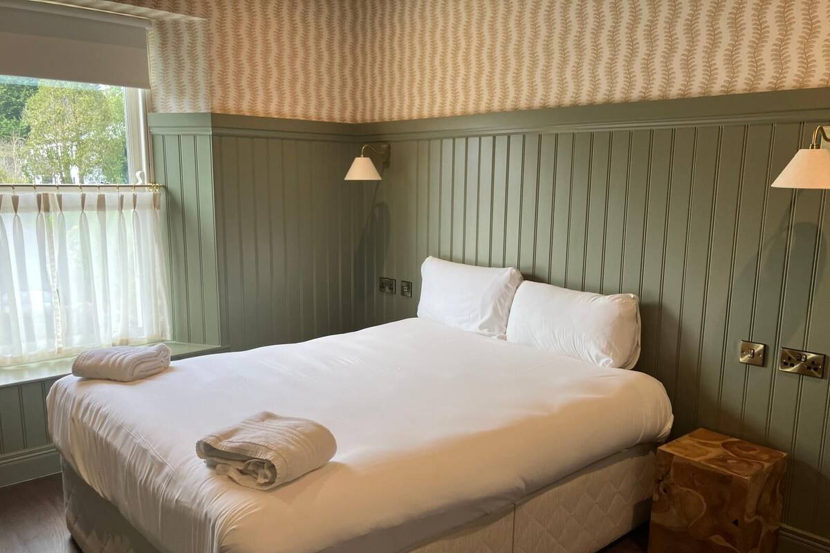 Cama bem arrumada em quarto com janela, luminária de cabeceira e mesinha de cabeceira de madeira.