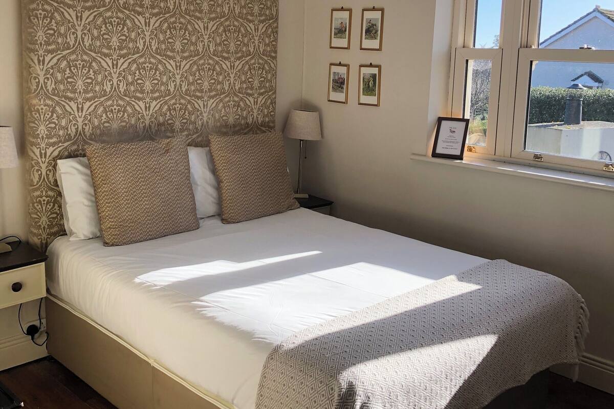 Uma cama bem arrumada com cabeceira estampada, mesa de cabeceira com abajur e janela com cortinas.