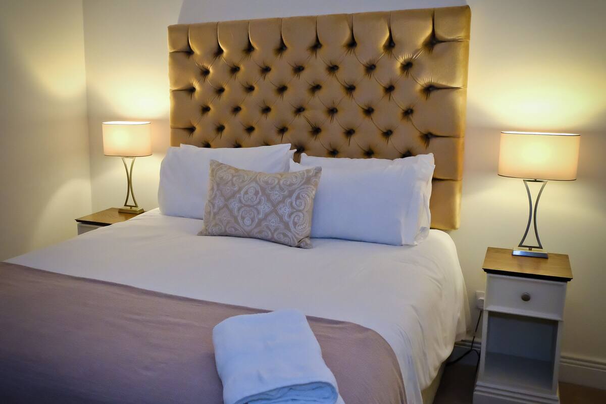 Uma cama bem arrumada com cabeceira estofada, duas mesas de cabeceira com abajures e uma toalha dobrada sobre a cama.
