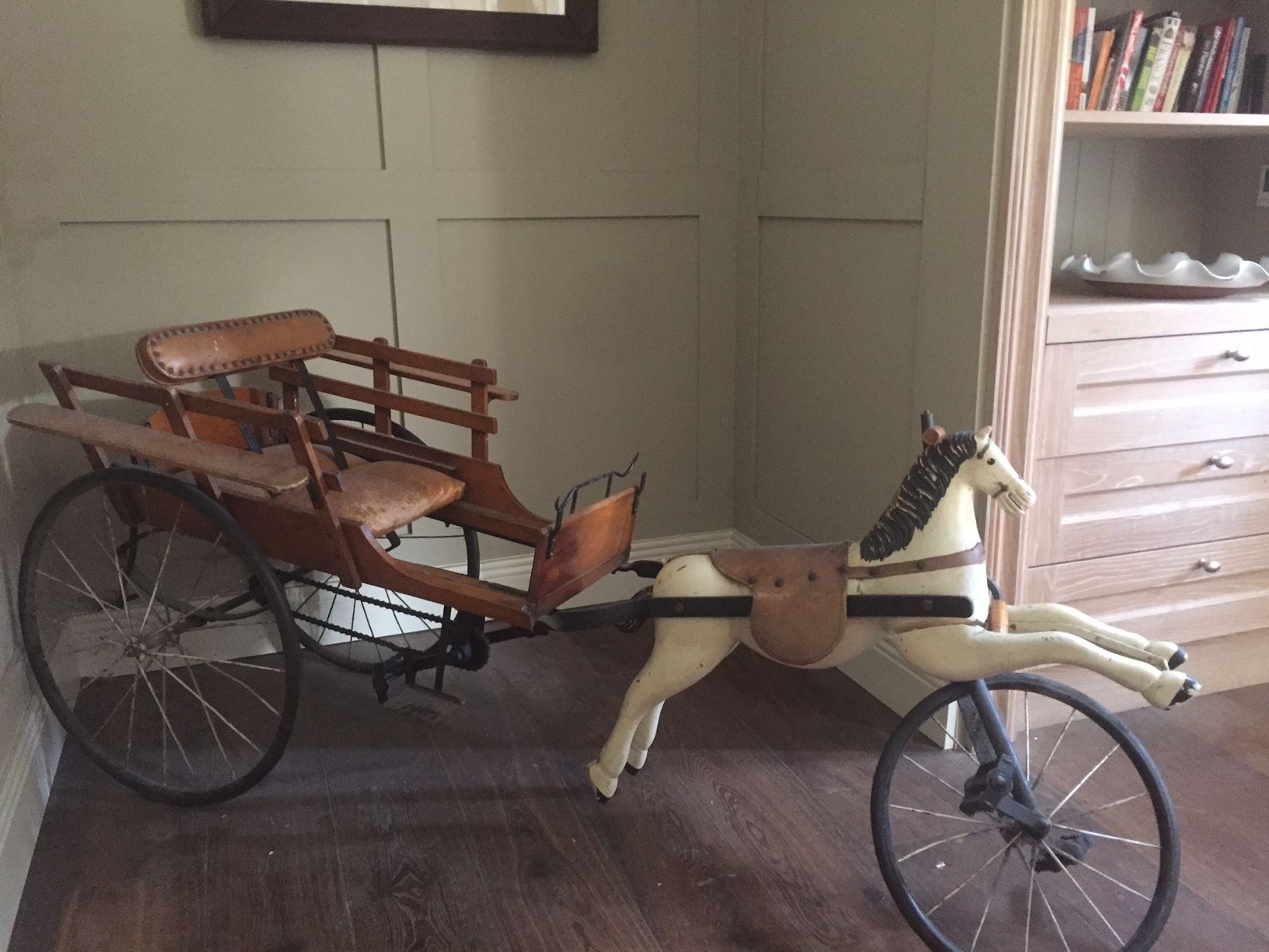 Un carruaje de madera con una figurina de caballo adherida, colocado en una habitación con una estantería y un aparador al fondo.