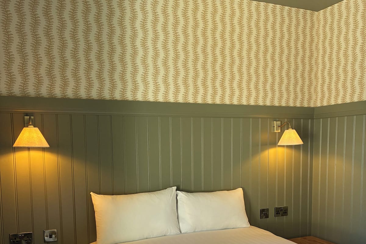 Cama bem arrumada com duas luminárias de cabeceira, mesa de cabeceira de madeira e parede com painéis verticais.
