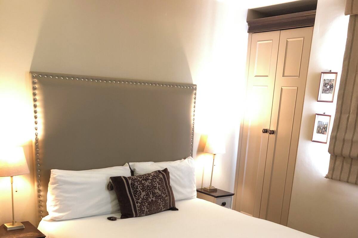 Uma cama bem arrumada com cabeceira, duas mesas de cabeceira com abajures, piso de madeira e um guarda-roupa com espelho.