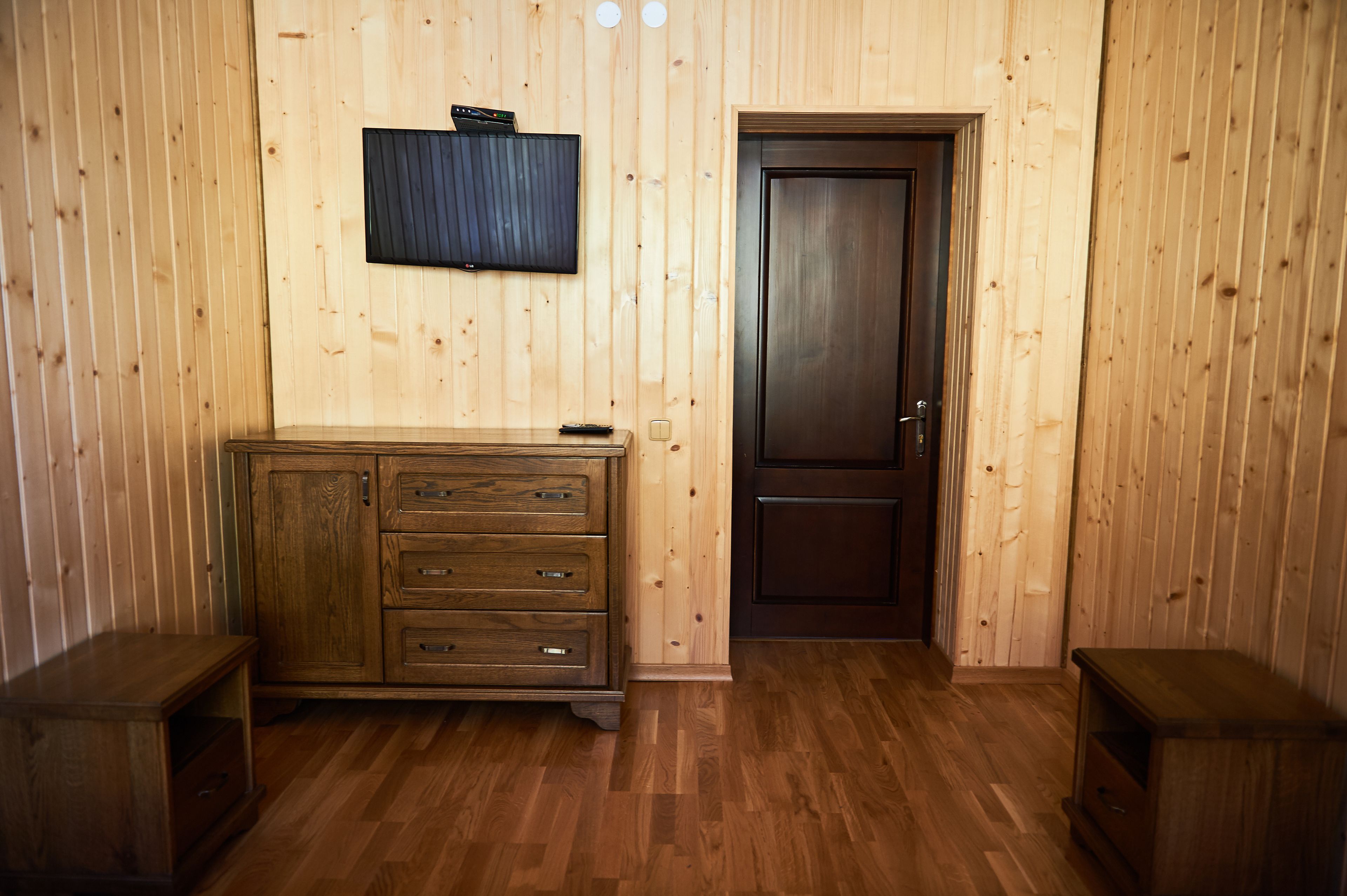 A room with wooden walls and flooring, a wooden cabinet with drawers, a television mounted on the wall, and a door.