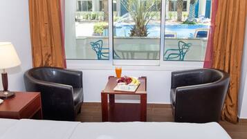 A hotel room with a bed, two armchairs, a small table with fruit and juice, and a window with a view of a pool and palm trees.