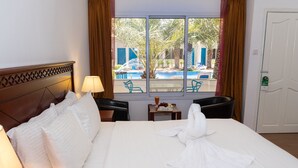Una habitación de hotel con una cama, una mesa con una lámpara, una silla, una ventana con vista a una piscina y palmeras, y una puerta.