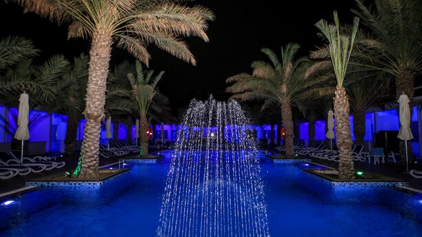 Una piscina iluminada con luz azul, con una fuente de agua, rodeada de palmeras y sillones de descanso.