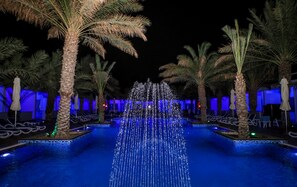 A nighttime swimming pool with a water fountain, surrounded by palm trees and lounge chairs under blue lighting. - Fujairah Hotel & Resort (Al-Fujairah)