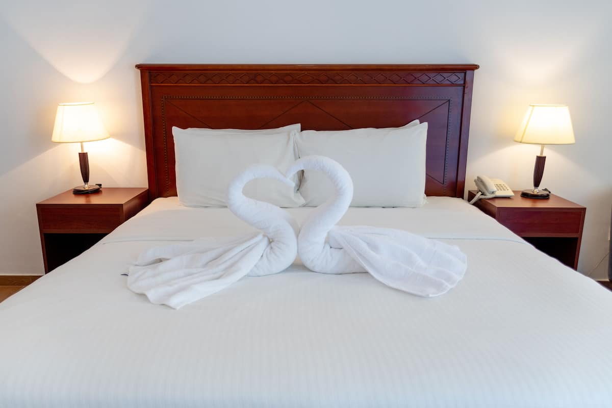 A neatly made bed with two towels arranged in a heart shape, a wooden headboard, and bedside lamps on either side.