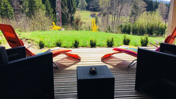Una terraza con sillas de mimbre y una mesita, con vista a un área verde con árboles y una lejana cadena montañosa.