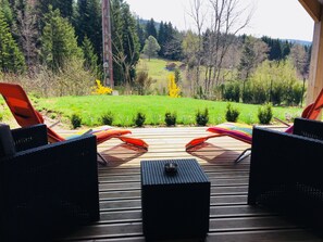 Una terraza con sillas de mimbre y una mesita, con vista a un área verde con árboles y una lejana cadena montañosa.