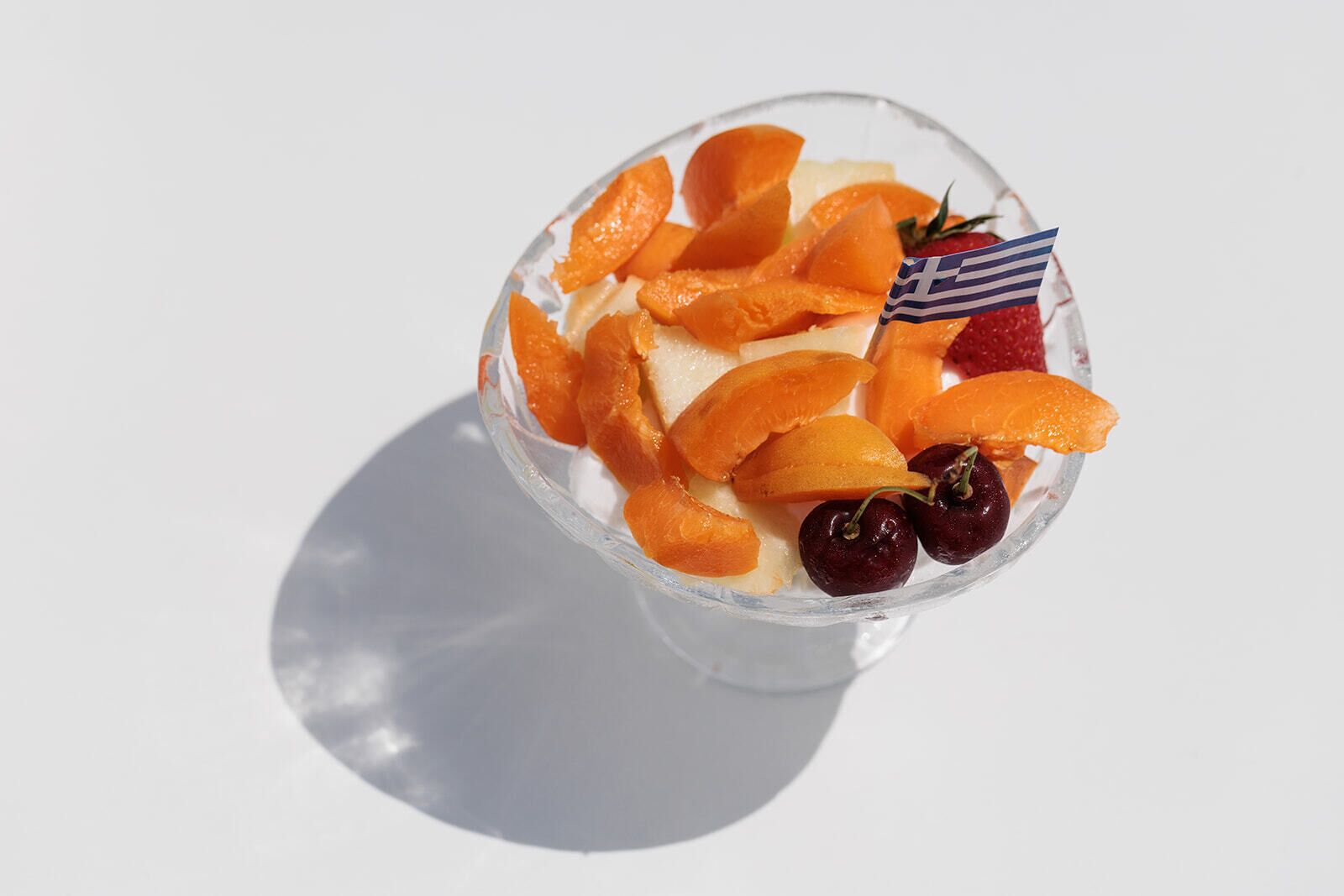 Un bol de vidrio transparente con rodajas de naranja, fresas y cerezas, con una pequeña bandera griega en la parte superior.