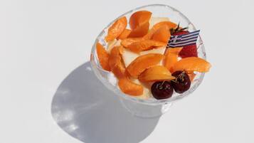 Un bol de vidrio transparente con rodajas de naranja, fresas y cerezas, con una pequeña bandera griega en la parte superior.