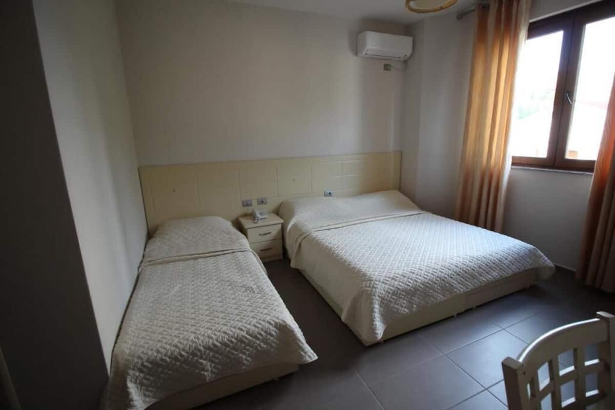 Quarto com duas camas, uma mesa de cabeceira, uma cadeira e uma janela com cortinas.