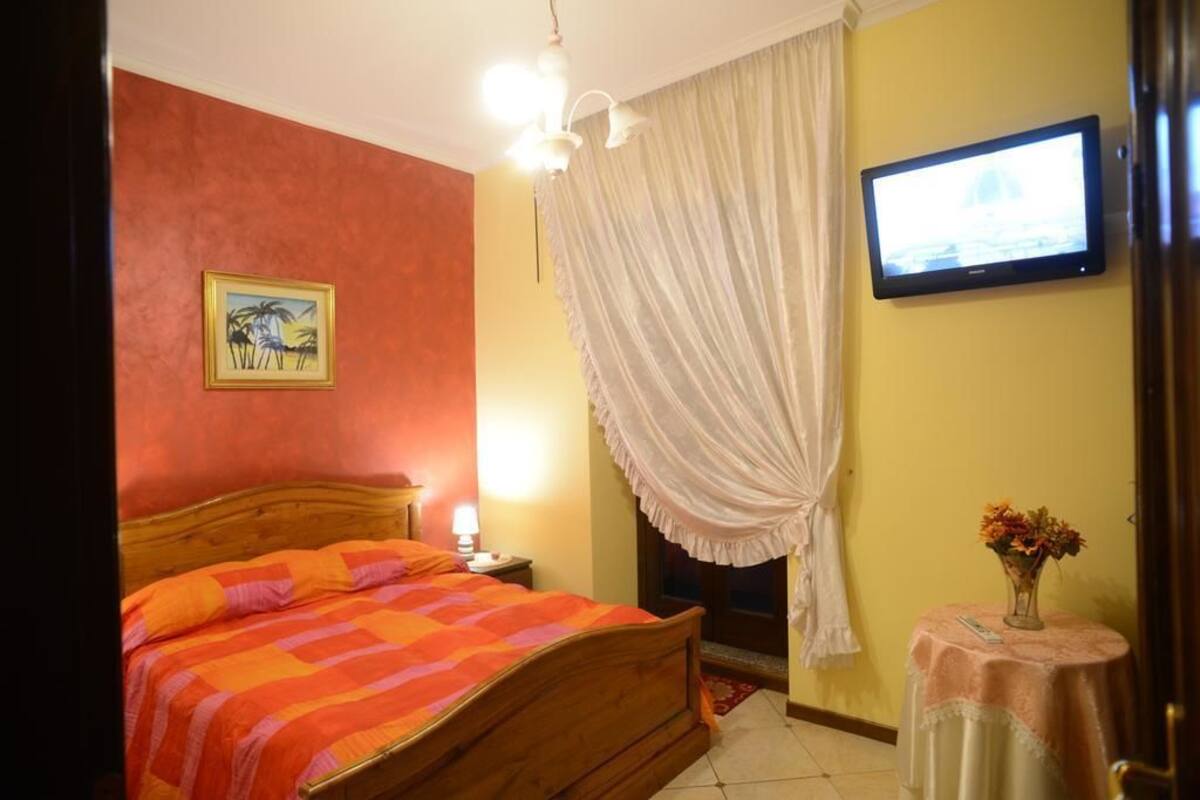 A hotel room with a bed, a television, a painting, a curtain, and a small table with a vase.