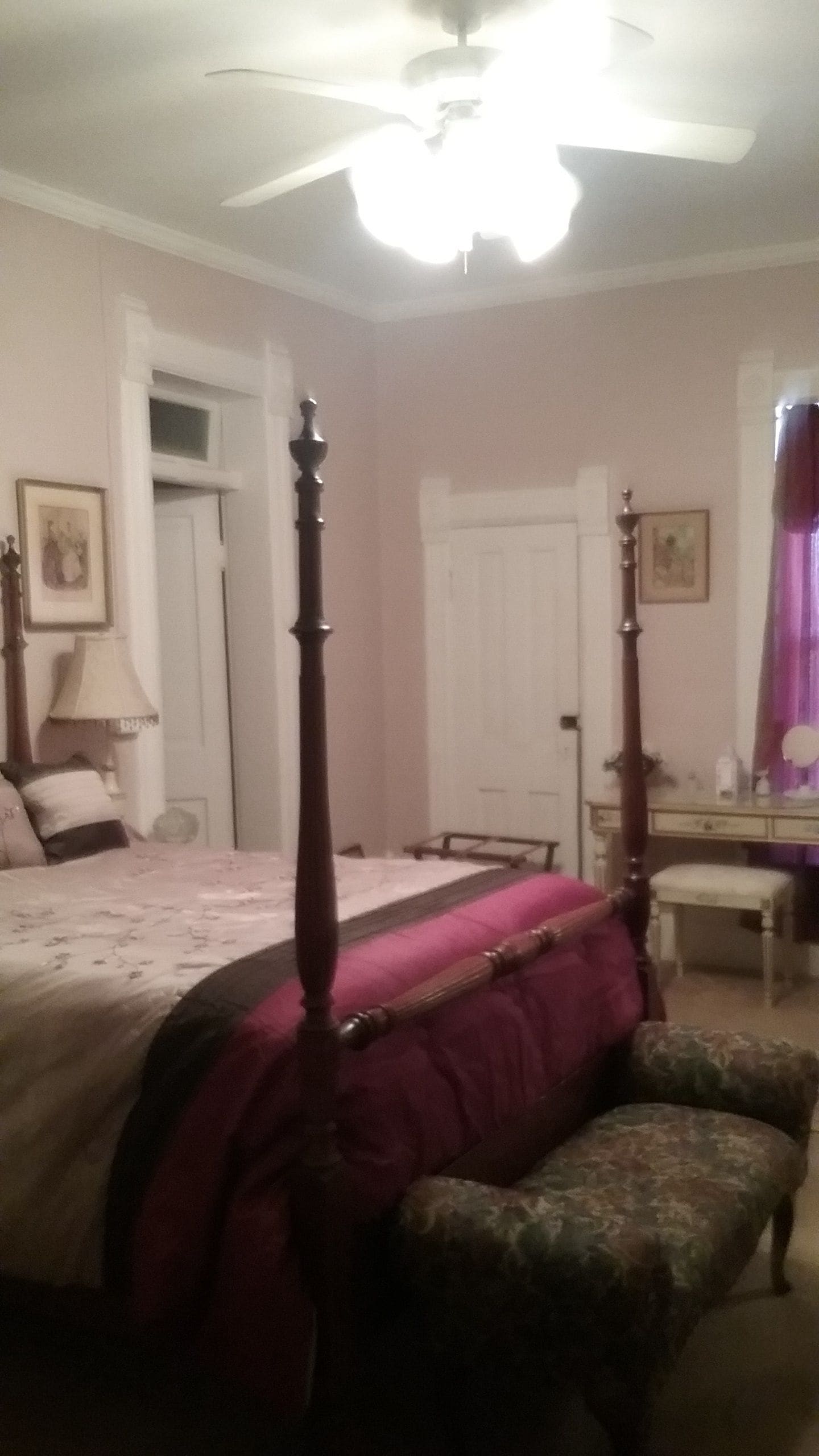 A bedroom with a four-poster bed, a wooden bench, a desk, and a ceiling fan.