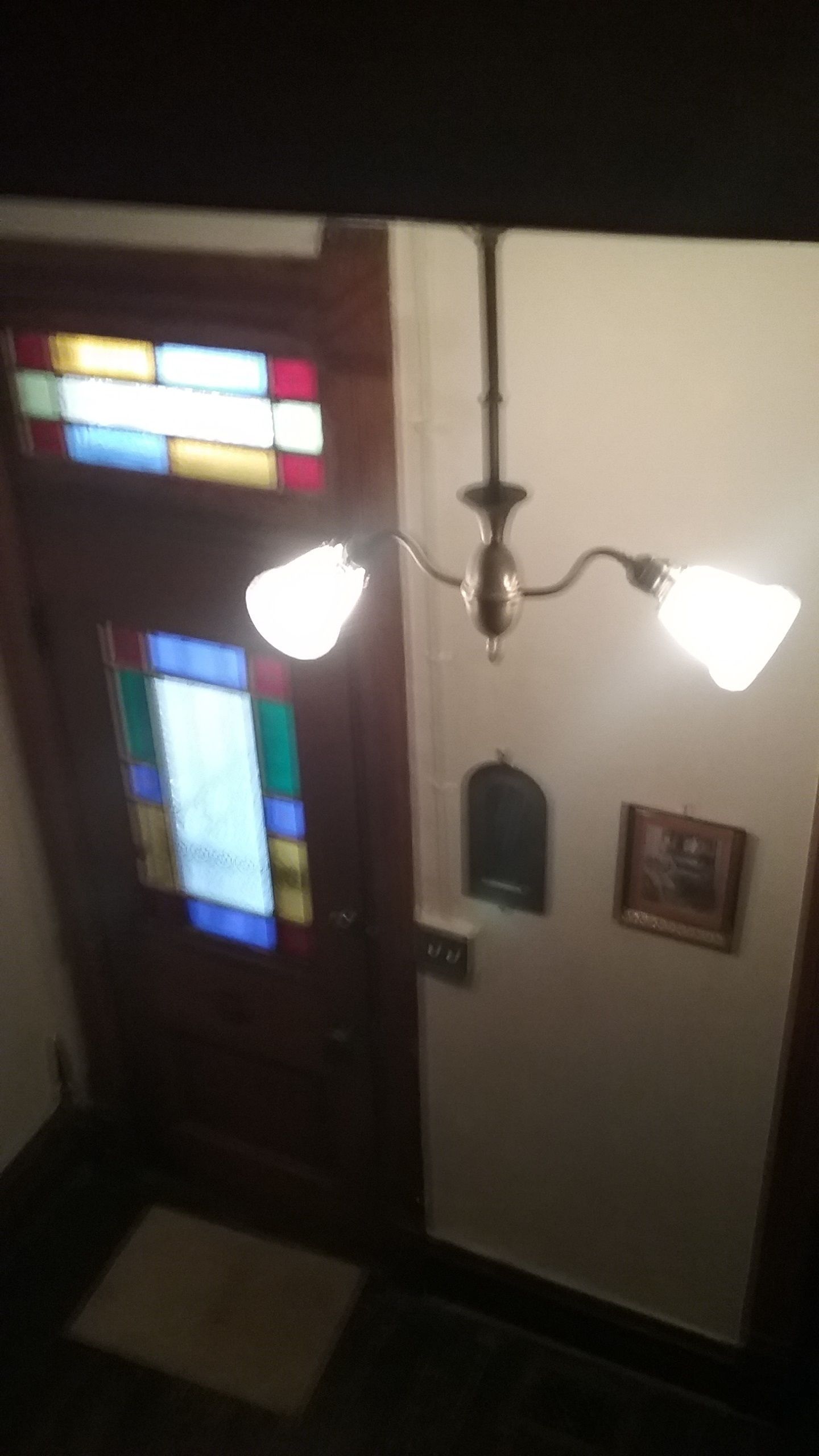 Un pasillo con una puerta de vitral, una lámpara de pared y un pequeño cuadro enmarcado.