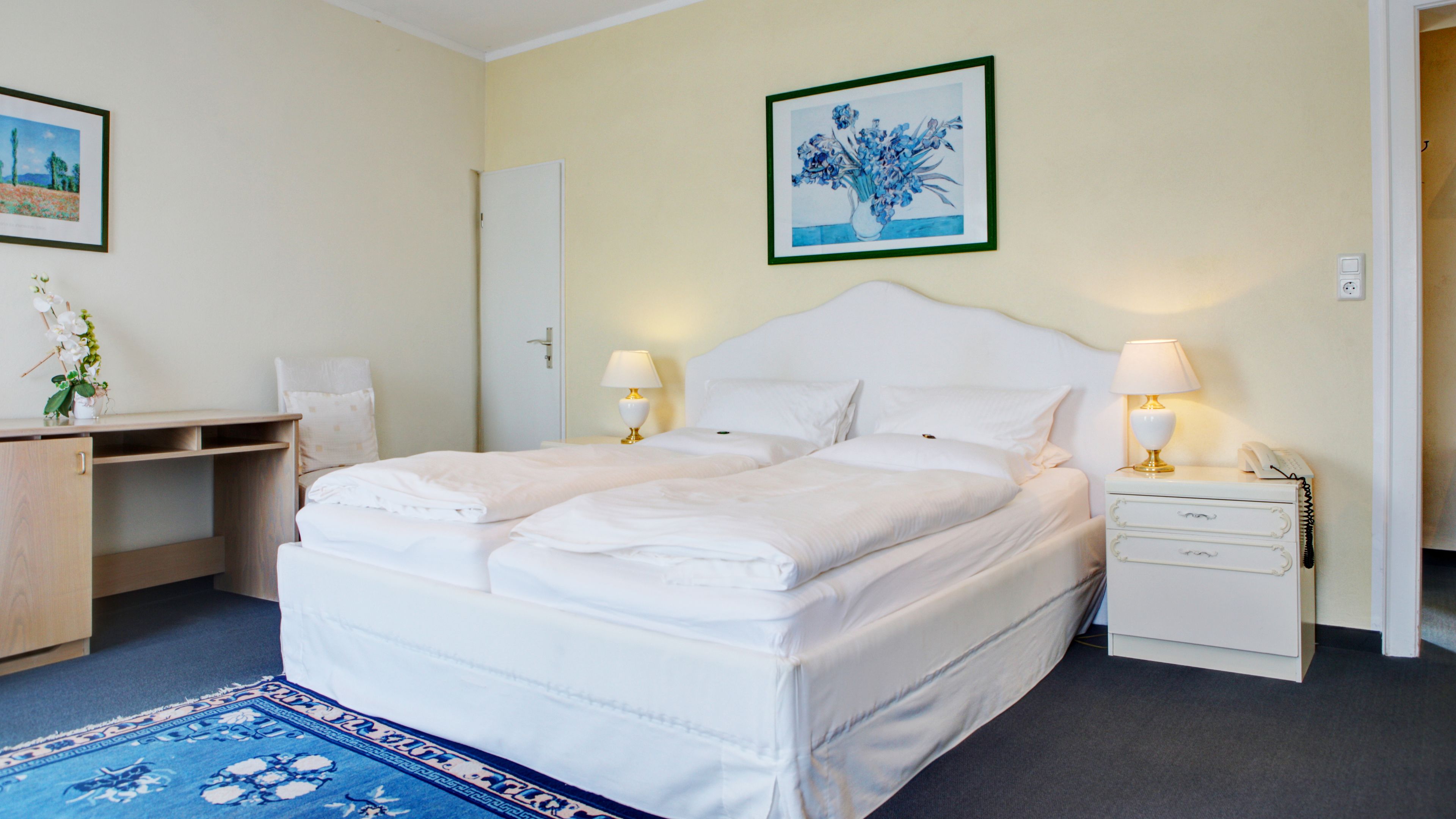 A double bed with white linens, a bedside table with a lamp, a nightstand with a phone, a desk with a chair, and a framed picture on the wall.